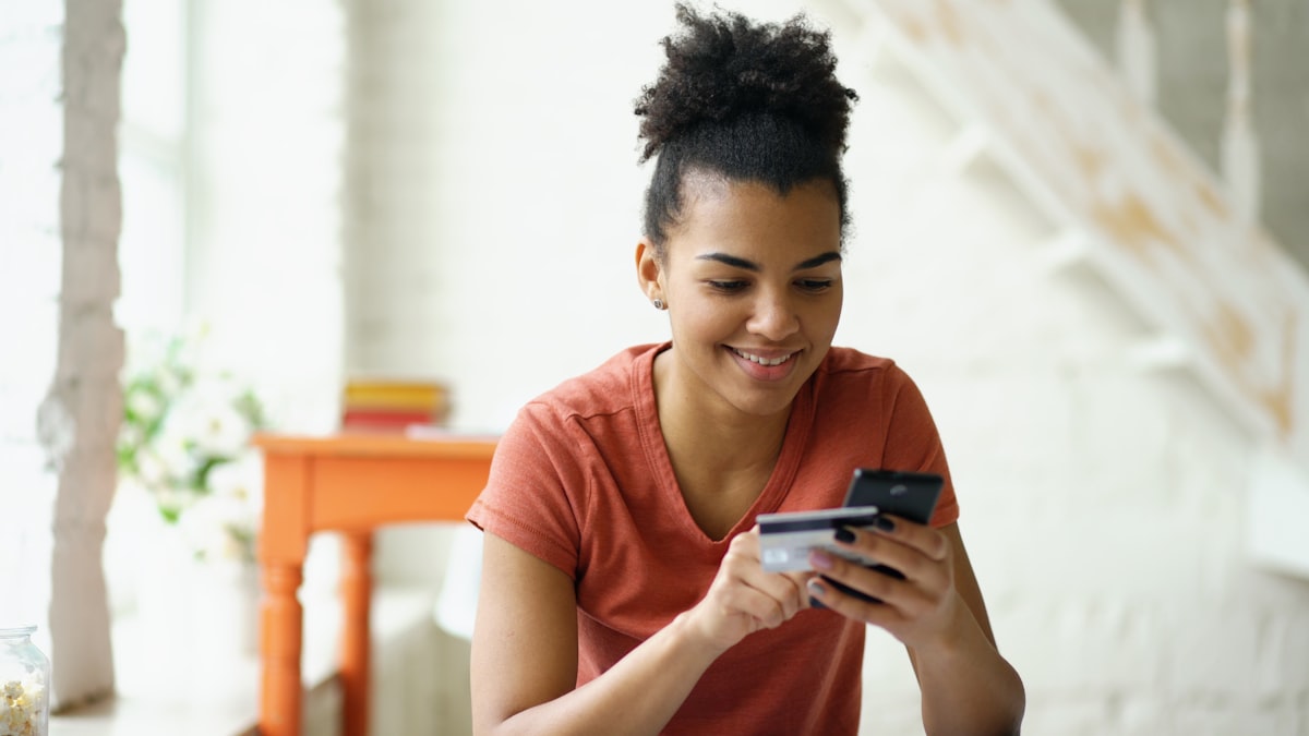 Mulher sorrindo usando smartphone e segurando cartão de crédito — representando serviços financeiros digitais
